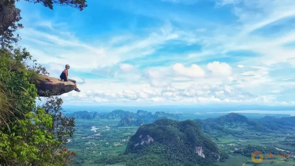 This image shows a traveler sitting on a cliff ledge at Khao Ngon Nak viewpoint in Krabi, Thailand, overlooking mountains, jungle and the Andaman Sea, highlighting hiking and scenic viewpoints as one of the best things to do in Krabi.