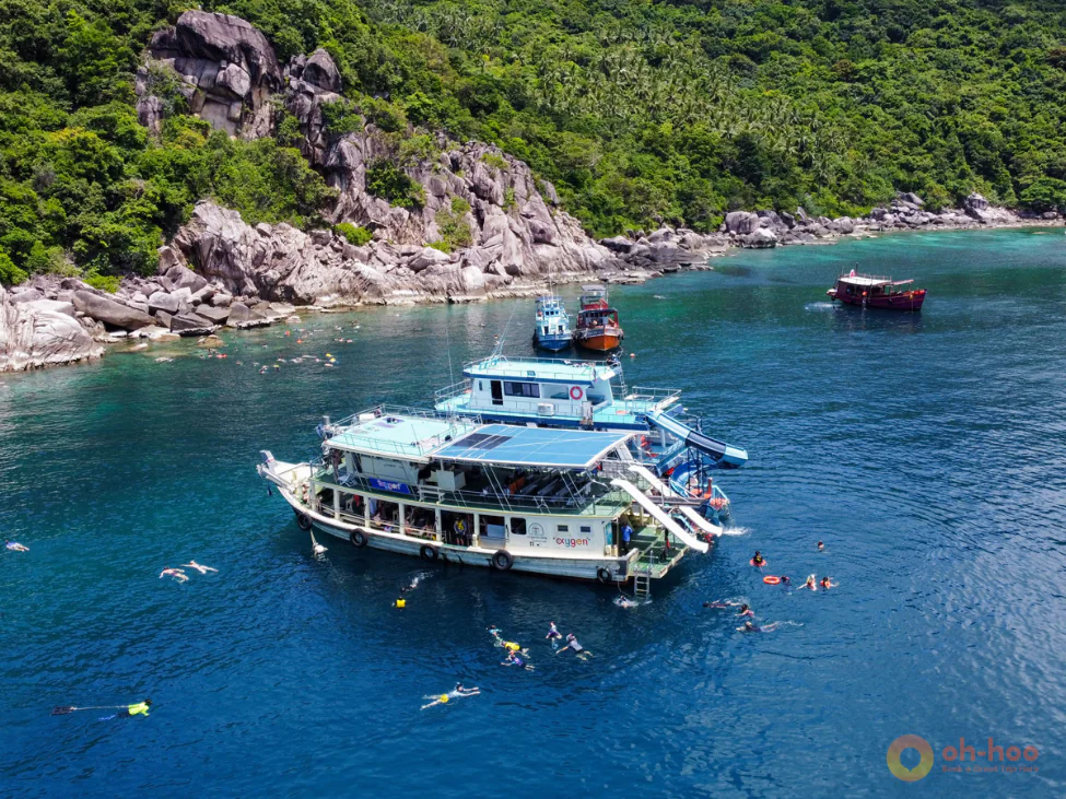Aerial view of tour boats in clear turquoise water near Koh Tao, with visible coral patches and snorkelers exploring the bay.