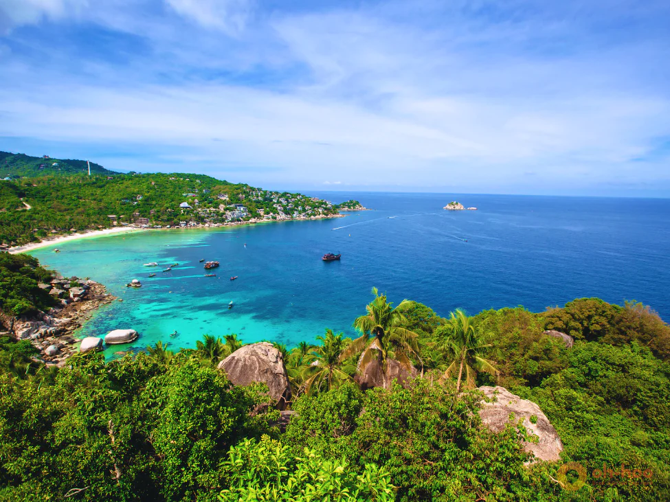 Aerial view of a hidden bay in Koh Tao with turquoise water, longtail boats, rocky shoreline, and tropical palm trees