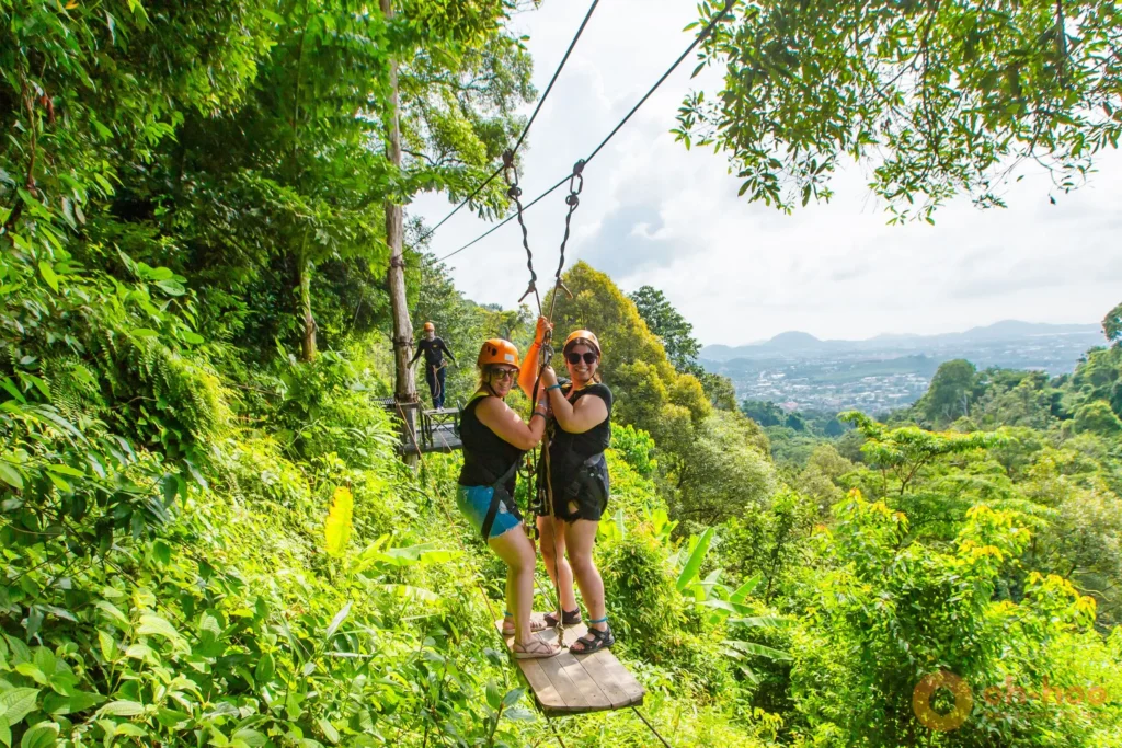 Two smiling women in helmets and harnesses on a Phuket zipline platform high above the jungle canopy