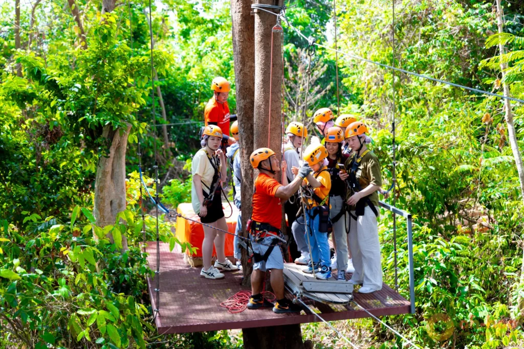 A trained guide fitting a child's safety harness before a zipline ride, with helmets and belay gear on a jungle platform