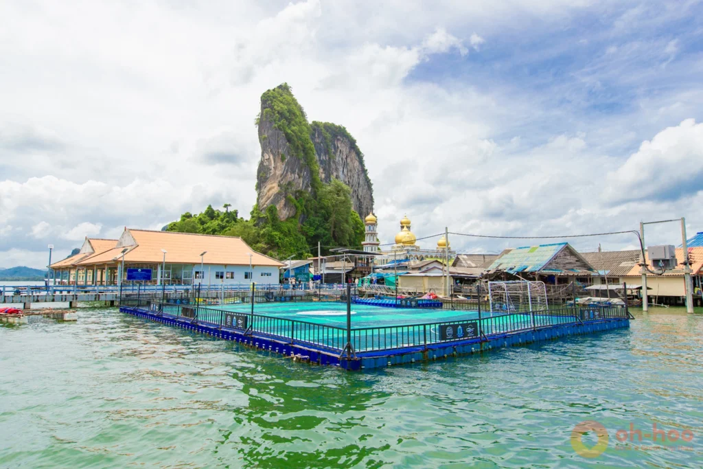 The floating village of Koh Panyee featuring its famous floating football pitch, golden mosque domes, and towering limestone cliff in Phang Nga Bay.