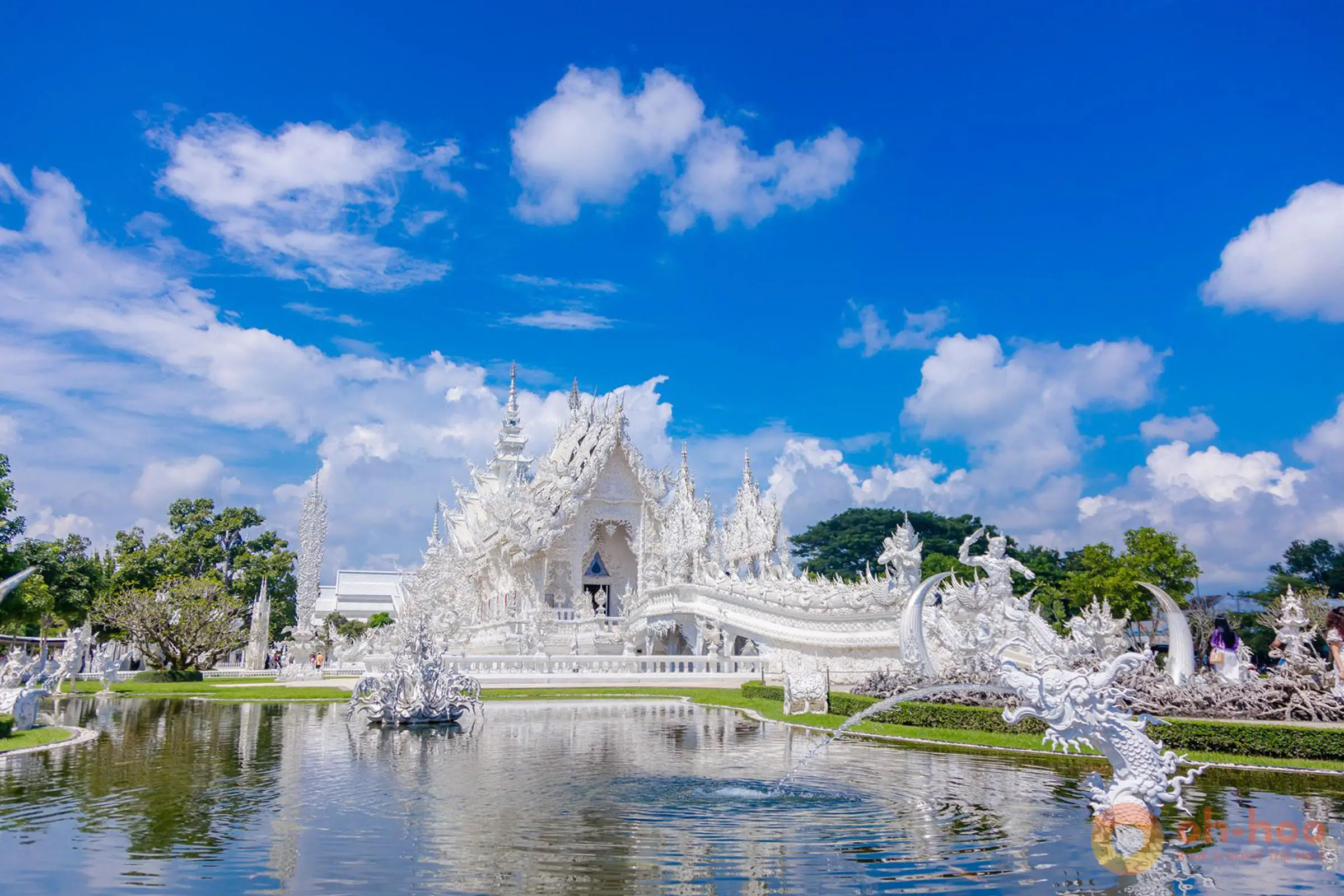 The view of the gleaming White Temple (Wat Rong Khun) reflected in the surrounding pond under a clear blue sky with fluffy clouds in Chiang Rai.