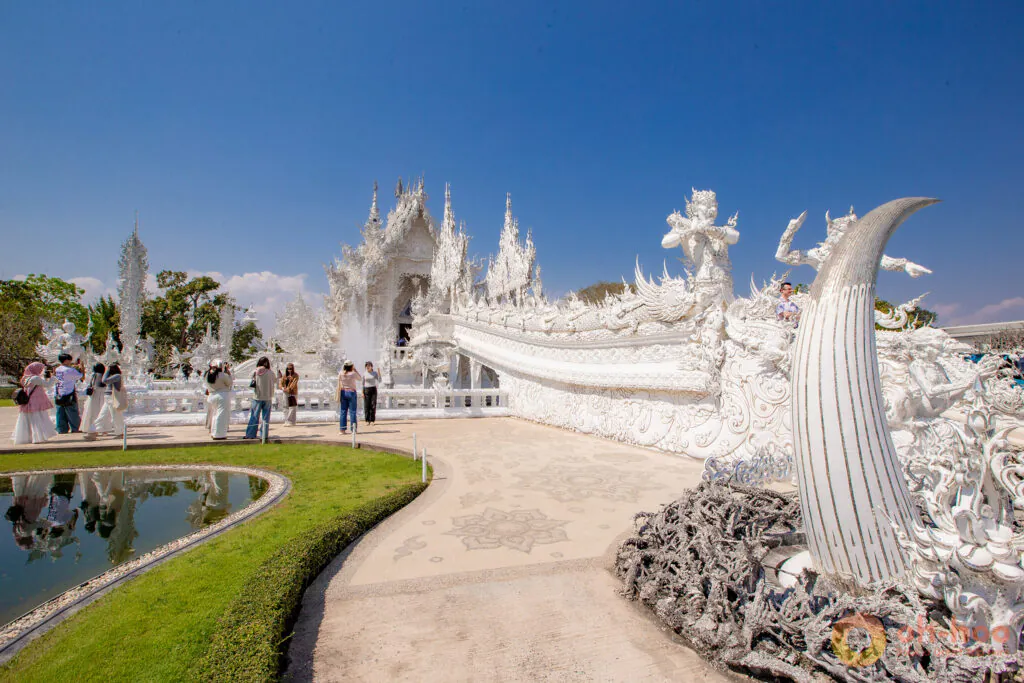 Visitors walking along the paved path past large white sculptures and the Bridge of Rebirth toward the main hall at Wat Rong Khun.