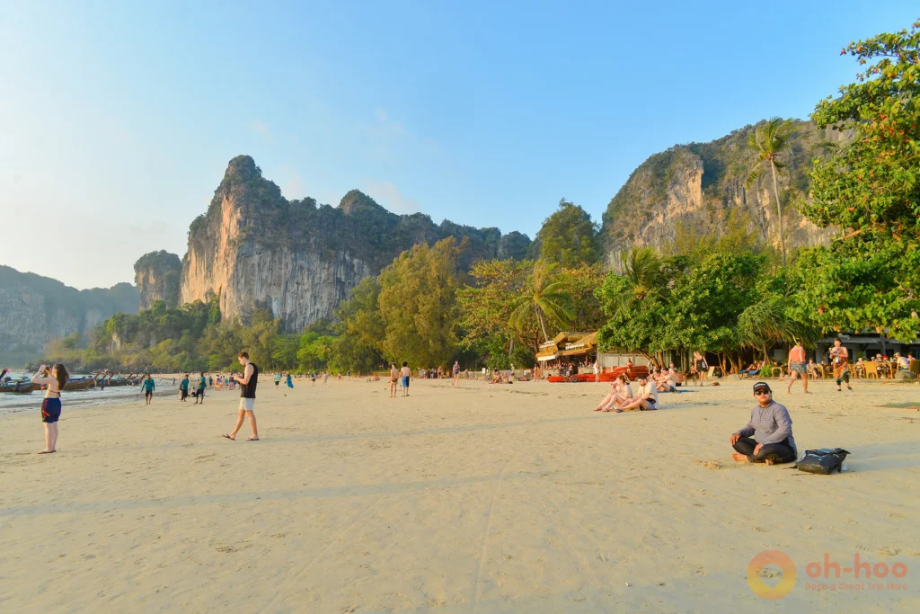 A wide-angle panoramic view of Railay West beach featuring turquoise waters, towering limestone cliffs, and longtail boats anchored along the shore.