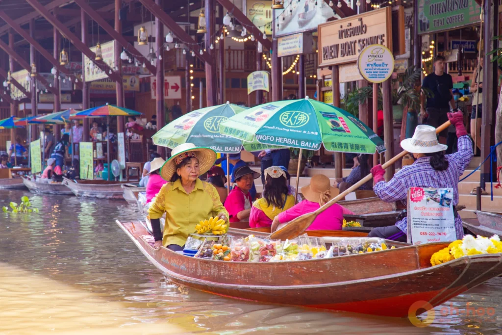 A woman paddling a wooden sampan carrying visitors past fruit vendors at Damnoen Saduak