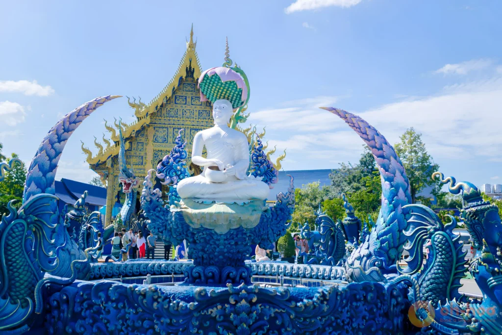 White Buddha seated on a lotus among blue and gold naga sculptures in front of the Blue Temple hall in Chiang Rai