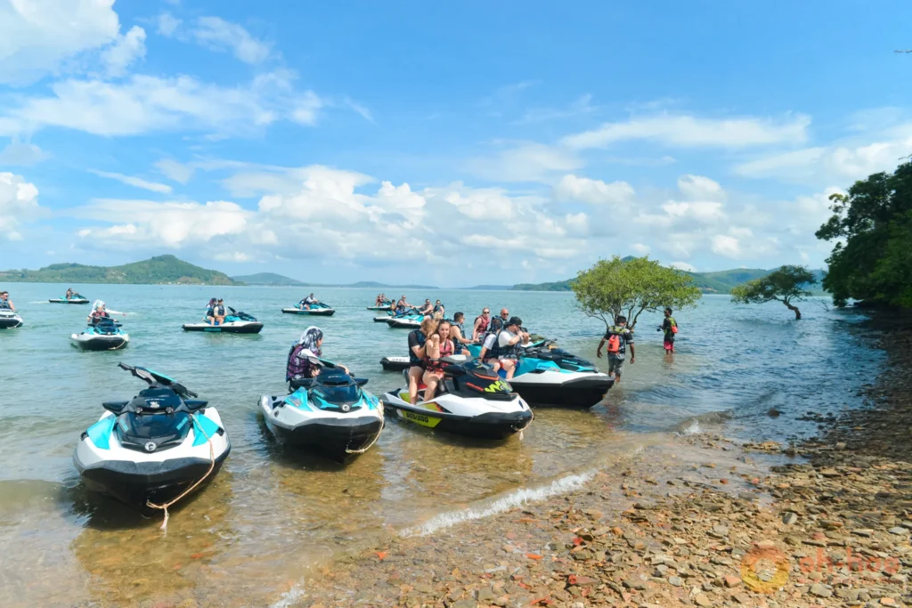 Row of jet skis lined up on the sand at a busy Phuket beach with operators waiting