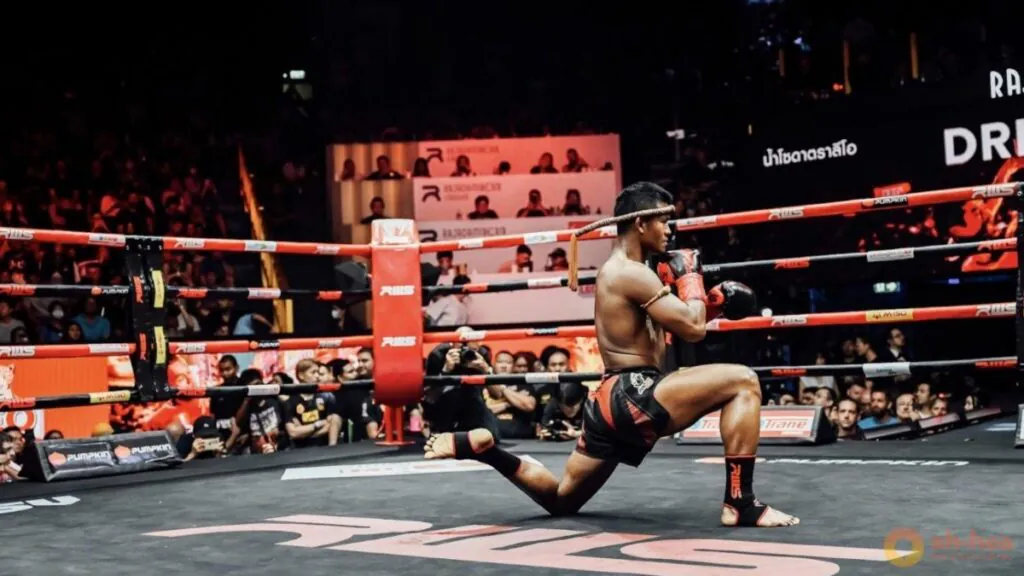 A fighter kneels on the canvas during a pre-fight ritual inside a ring, wearing a mongkhon headband and gloves.