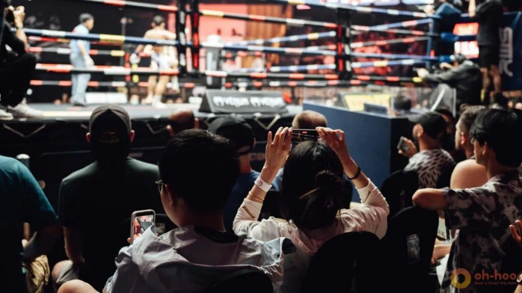 Spectators sit ringside, watching fighters in the illuminated canvas ring during a muay thai fight bangkok.