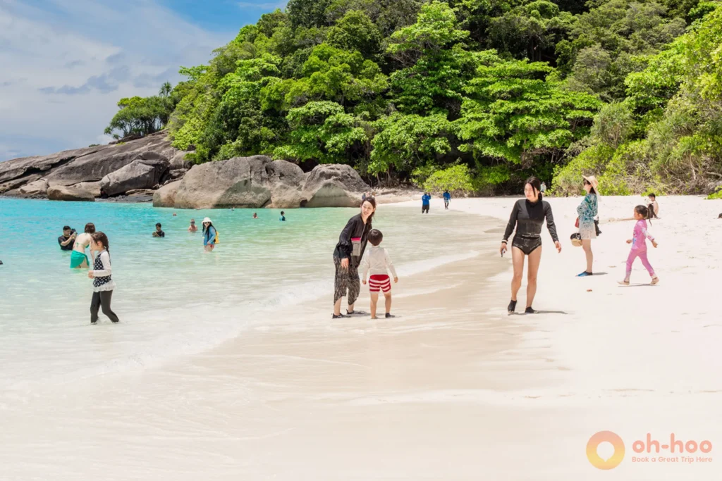 Visitors walking and relaxing on a white sand beach at the Similan Islands with a lush green hill in the background
